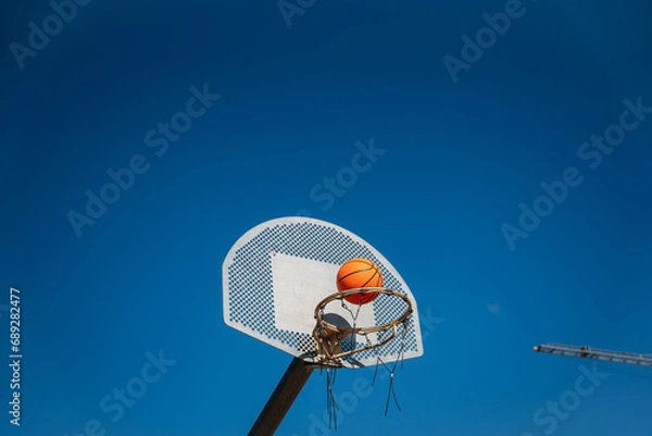 Fototapeta Basketball basket seen from below and one side while an orange ball is falling from above. Contrasted with a blue sky on a sunny, clear day.