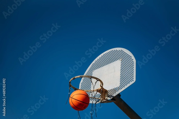 Fototapeta Basketball basket seen from below and one side while an orange ball is falling from above. Contrasted with a blue sky on a sunny, clear day.