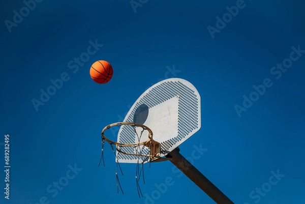 Obraz Basketball basket seen from below and one side while an orange ball is falling from above. Contrasted with a blue sky on a sunny, clear day.