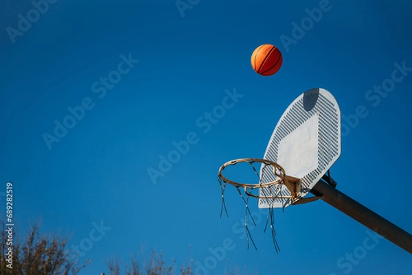 Fototapeta Basketball basket seen from below and one side while an orange ball is falling from above. Contrasted with a blue sky on a sunny, clear day.
