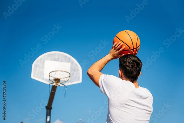 Fototapeta Young Caucasian boy with his back turned, wearing a white t-shirt ready to throw a basketball at a metal basket and the clear blue sky on a sunny day.