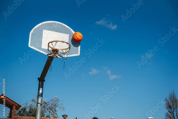 Fototapeta Basketball basket seen from below and one side while an orange ball is falling from above. Contrasted with a blue sky on a sunny, clear day.