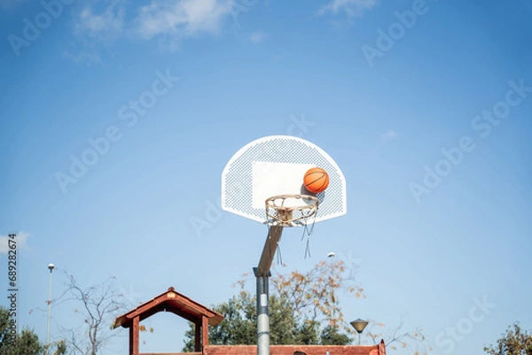 Fototapeta Basketball basket seen from below and one side while an orange ball is falling from above. Contrasted with a blue sky on a sunny, clear day.