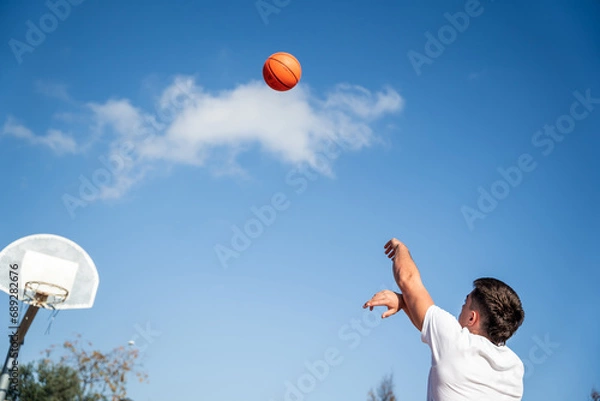 Obraz Young Caucasian boy with his back turned, wearing a white t-shirt throwing a basketball at a metal basket and the clear blue sky on a sunny day.