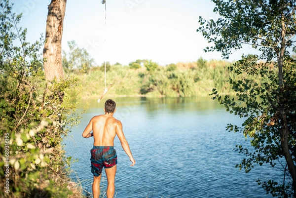 Obraz Unrecognizable muscular young guy seen from behind thinking about jumping into water from a rope/zip line. Caucasian boy doing water sport.