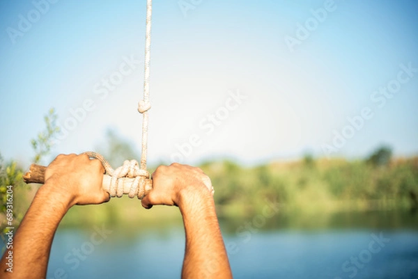 Obraz Detail of hands of young Caucasian man grabbing a stick and a rope tied to a tree to jump into the water in a lake. Sunny summer day.