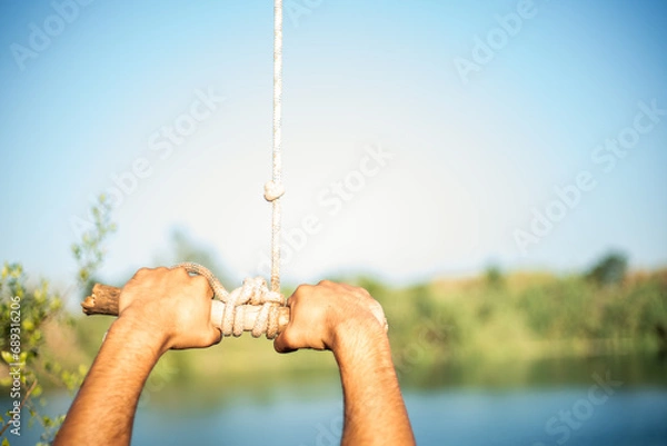 Fototapeta Detail of hands of young Caucasian man grabbing a stick and a rope tied to a tree to jump into the water in a lake. Sunny summer day.