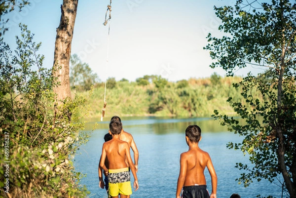 Obraz A group of unrecognizable young boys seen from behind thinking about jumping with a rope into the water of a lake in summer.