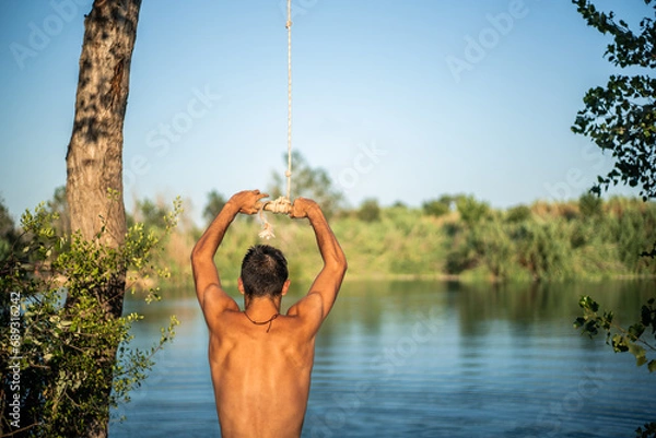Fototapeta Unrecognizable muscular young guy seen from behind thinking about jumping into water from a rope/zip line. Caucasian boy doing water sport.