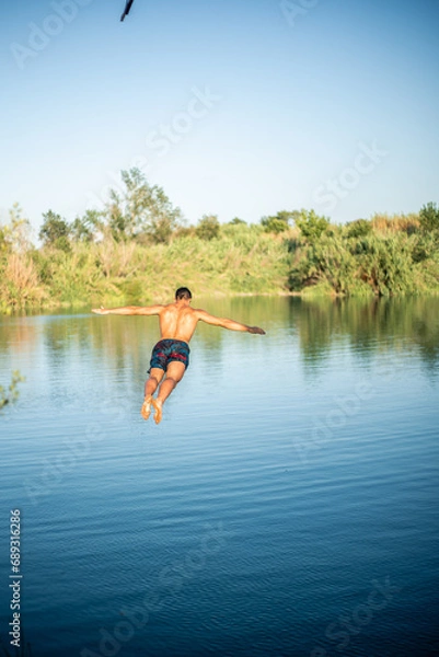 Obraz Unrecognizable muscular young guy seen from behind jumping into the water from a rope/zip line while doing a flip. Caucasian boy doing water sport.