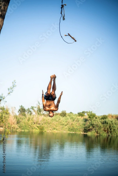 Fototapeta Unrecognizable muscular young guy seen from behind jumping into the water from a rope/zip line while doing a flip. Caucasian boy doing water sport.