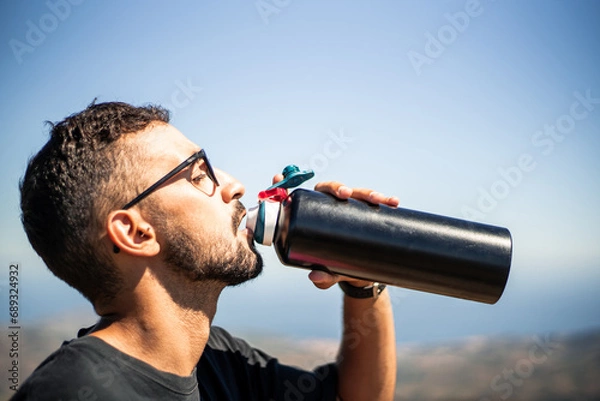 Fototapeta Young Caucasian man of North African nationality thirstily drinking water from a black unbranded canteen or bottle on a very sunny and hot summer day. Profile view.