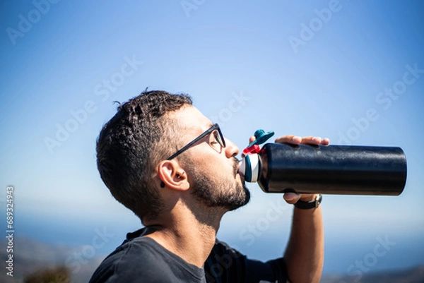 Fototapeta Young Caucasian man of North African nationality thirstily drinking water from a black unbranded canteen or bottle on a very sunny and hot summer day. Profile view.