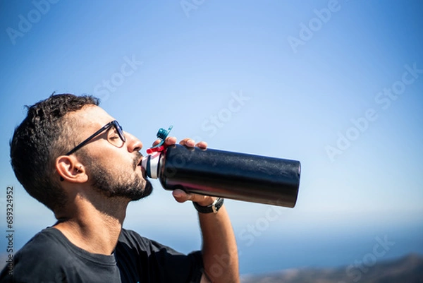 Fototapeta Young Caucasian man of North African nationality thirstily drinking water from a black unbranded canteen or bottle on a very sunny and hot summer day. Profile view.