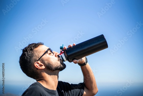Fototapeta Young Caucasian man of North African nationality thirstily drinking water from a black unbranded canteen or bottle on a very sunny and hot summer day. Profile view.