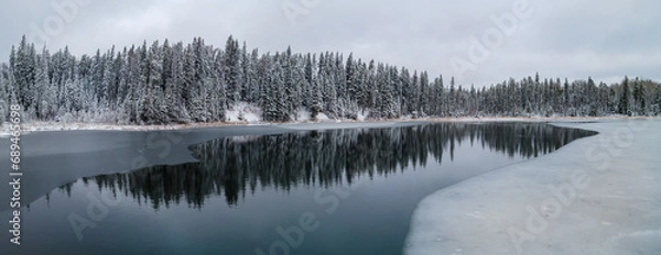 Fototapeta A panoramic view of a cold looking winter scene at a partially frozen lake that is surrounded by an evergreen forest. The trees are covered with heavy snow.  Tree reflections can be seen in the calm w