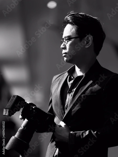 Fototapeta Vertical black and white portrait of an Asian man with short, cool black hair, wearing glasses, wearing a black suit, business man, holding a camera. with a background of light and shadow