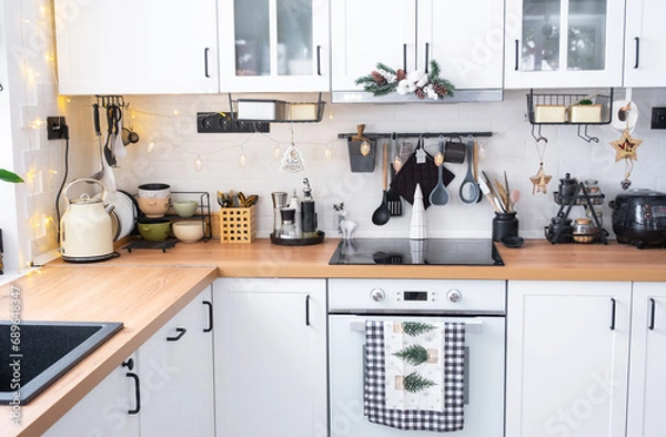 Fototapeta White Christmas decor in white kitchen, towel with Christmas trees, a branch of a fir tree on cabinets, induction cooker with oven. New Year, Christmas mood, cozy home. 