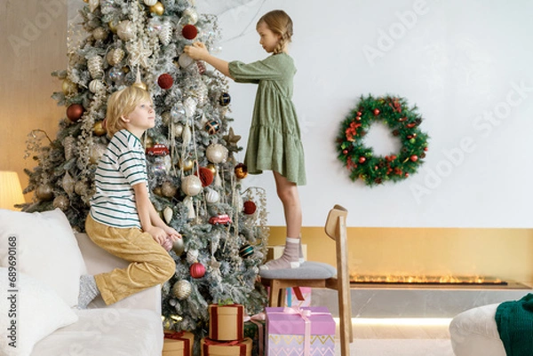 Obraz boy and girl decorate Christmas tree by balls and have fun at home. sister stands on chair and brother its on couch.