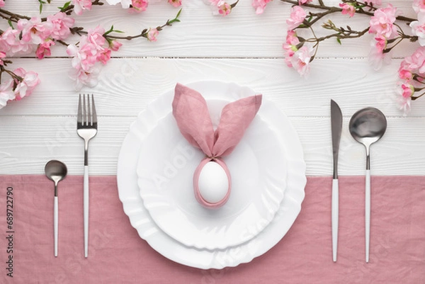 Fototapeta Easter lunch or brunch invitation. Festive table setting with pink spring flowers, cutlery. Bunny made of linen napkin and egg. Happy Easter holiday concept menu. Empty plate on white wood background.