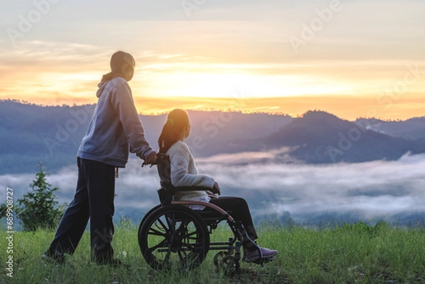 Obraz Silhouette of woman in wheelchair with care helper walking around in spring nature at sunrise misty mountains.