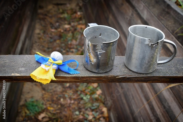 Fototapeta War in Ukraine. Belongings of Ukrainian warriors defending his country during the war such as iron mugs deformed by fragments of enemy shells and Ukrainian traditional angel doll as amulet from death.