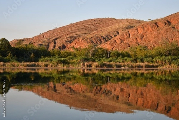 Fototapeta Hills of red earth and red rocks in the Kimberley area of Western Australia rise beside a river. A forest of grasses and trees border the hills. Both are reflected in the water. The sky is blue.