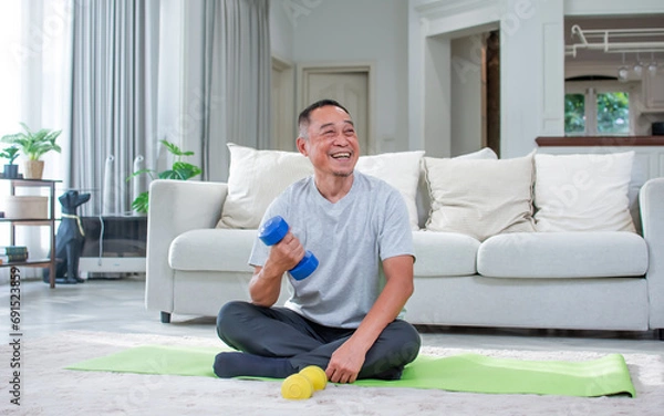 Fototapeta Asian aging senior old man wearing casual comfortable t shirt, sitting on floor in living room at cozy home, doing exercise with happiness, smiling. Healthcare, Retirement Concept.