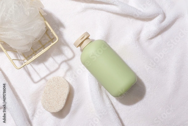 Fototapeta A bath sponge in an iron basket, a bar of soap and an unlabeled bottle of shower gel are displayed on a white fabric background. Minimalist space for advertising.