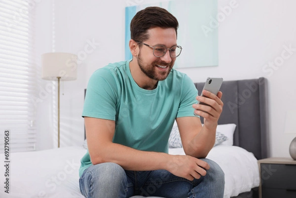 Fototapeta Happy man having video chat via smartphone in bedroom