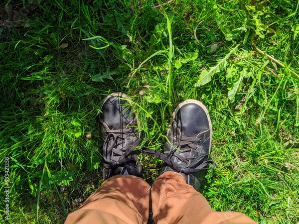 Obraz First person view of a man looking down at the shoes that are standing in the grass in the summer during the daytime.