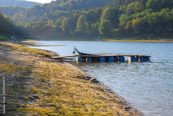 Fototapeta View of the wooden bridge on barrels on Lake Solina. There's a forest all around. People on the shore. Summer in the Bieszczady Mountains, Poland.