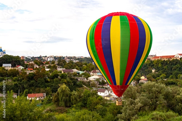 Obraz Multi colored balloon flying high in the sky over the city with a lot of trees. Balloon basket with people. Romantic trip. Copy space