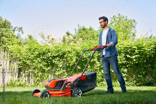 Fototapeta Handsome male gardener in gloves trimming turf with electric mower in garden at day. Side view of smiling bearded guy in denim outfit using lawn trimmer, while working outdoors. Concept of gardening.