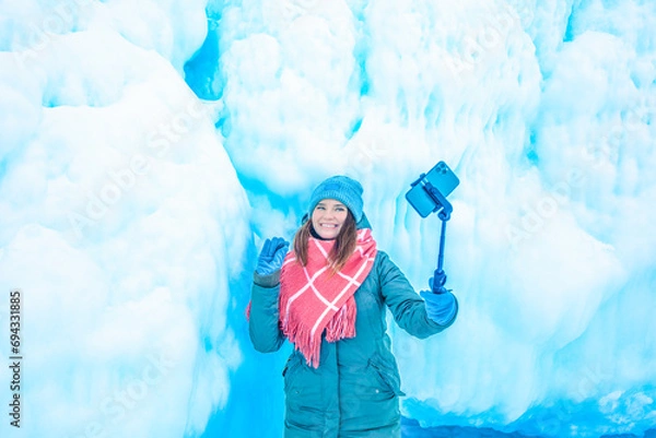 Obraz Cheerful female wonderer waves hand and talking via smartphone during video call while standing against big ice rock during winter vacation abroad 