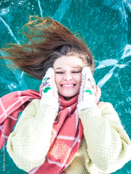 Obraz Portrait of a happy smiling female in cool mittens lying on a frozen Baikal lake with beautiful cracked ice sheets inside in winter day 