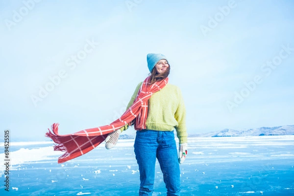 Obraz Gorgeous smiling female tourist with long stylish scarf enjoying warm winter day while standing on frozen Baikal lake during her trip to Siberia 