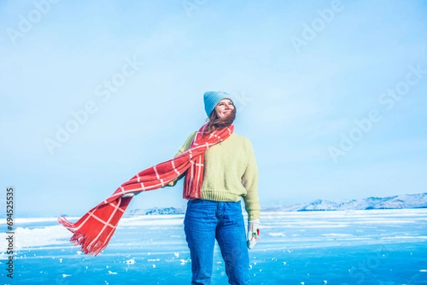 Obraz Gorgeous smiling female tourist with long stylish scarf enjoying warm winter day while standing on frozen Baikal lake during her trip to Siberia 