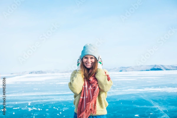 Obraz Cheerful female in stylish hat and mittens looking in camera while standing on frozen Baikal lake in sunny winter day 