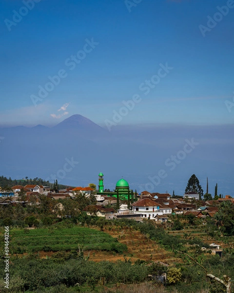 Fototapeta View of the mosque in a small village and the Semeru volcano. There are fields around the village. Sunny day. Batu. East Java. Indonesia.