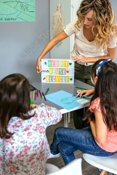 Fototapeta Female teacher explaining to students in classroom how it is a ecologic process while pointing to lightbox with text reduce, reuse, recycle. Sustainable lifestyle education concept.