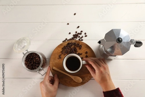 Fototapeta Man with cup of aromatic brewed coffee, top view. Moka pot, spoon and beans on white wooden table