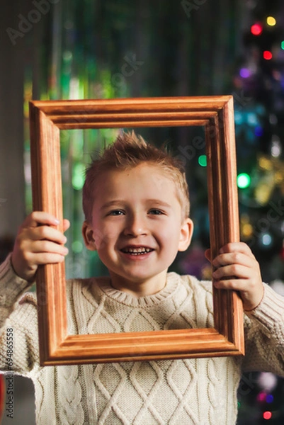 Obraz portrait of a cute boy of European appearance in a wooden picture frame against the backdrop of a Christmas tree in the room