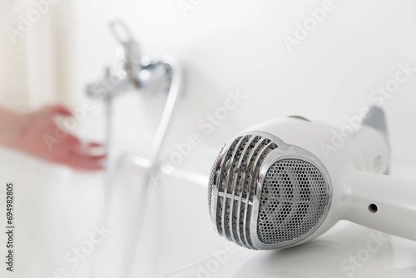 Fototapeta Hairdryer lying on a bathtub with hand and flowing water from a faucet in the background