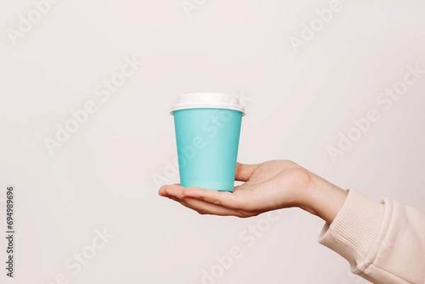 Obraz Blue eco paper cup with white lid with tea or coffee in a woman's hand isolated on a light background. Mockup with empty copy space for text or design. Recycling eco-friendly concept. Blank for logo