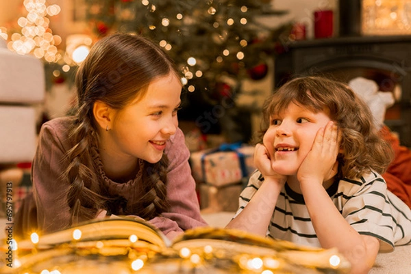 Obraz boy and girl have fun near Christmas tree, lay on floor reading a book near flashing lights, smiling and laughing during holidays