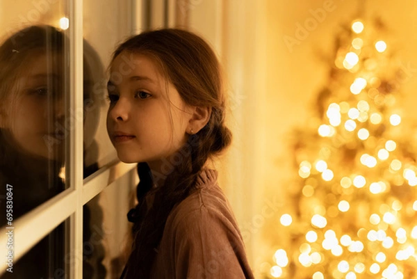 Obraz little girl with braids stand near window and look out, waiting santa claus with presents during christmas night