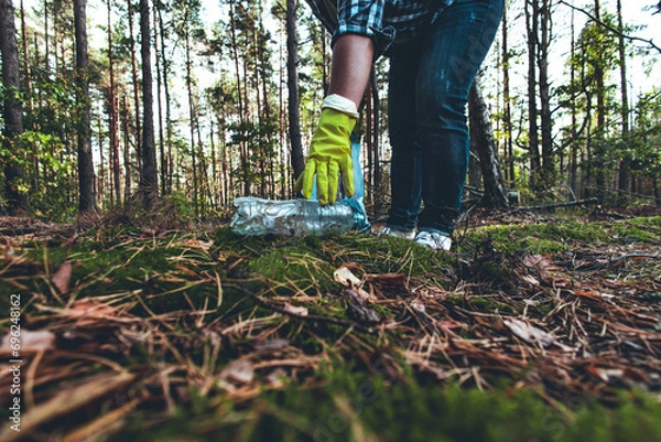 Obraz A plastic bottle lies on the moss in the sun. In the forest, a woman volunteer came up with a garbage bag to pick up plastic waste. Pollution of the forest with household waste and plastic garbage.
