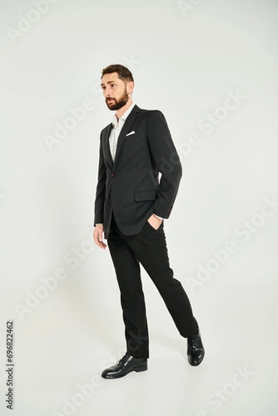 Fototapeta full length of wealthy businessman in black elegant suit posing with hand in pocket on grey