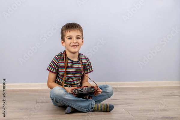 Obraz boy in a striped T-shirt sitting on the floor smiling with a retro camera in his hands on a blue background